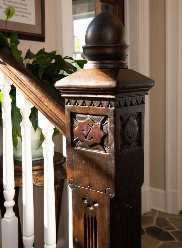 Close-up interior photograph of an ornate dark-stained timber newel post at the base of a staircase. The post features carved geometric and floral panels, decorative trim, and a rounded finial. White turned balusters, a timber handrail, neutral wall panelling, a framed artwork, indoor greenery, and irregular stone flooring are visible in the surrounding entry/stair area.