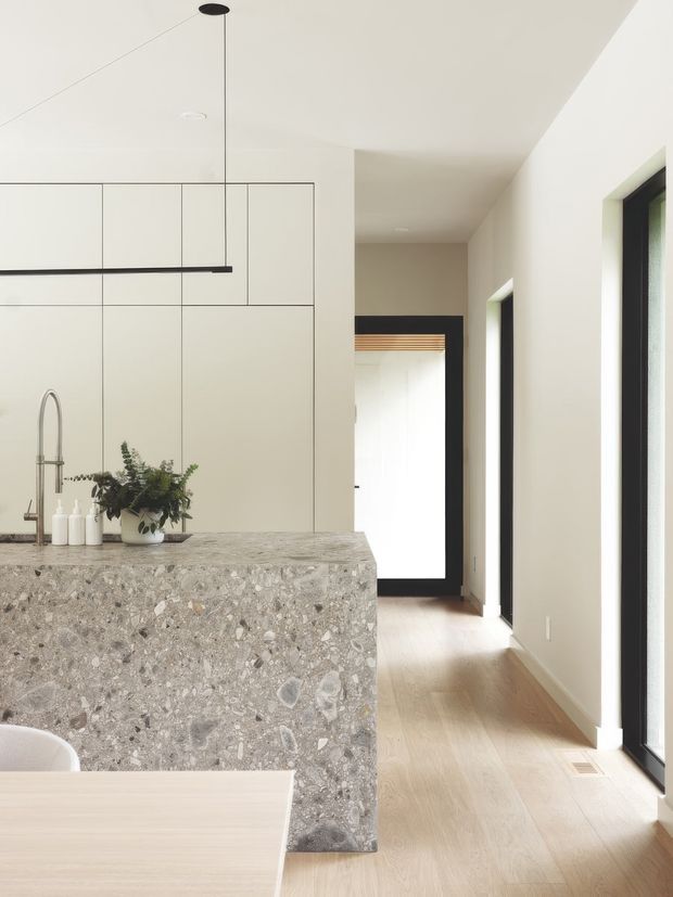 Bright, minimalist open-plan kitchen viewed toward a long hallway. The kitchen features full-height pale cabinetry, a substantial grey terrazzo island with integrated seating edge, a slim black linear pendant, and a stainless steel tap. Light timber flooring continues through the circulation space, which has tall black-framed windows and doors, white walls, and a dark-framed doorway at the end.