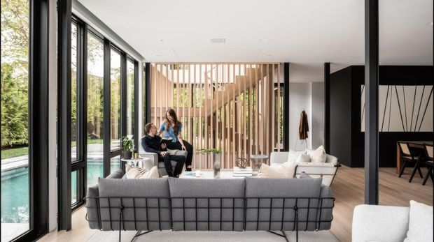 A bright, spacious contemporary living room with a large grey sectional sofa facing a light seating area. Floor-to-ceiling black-framed glazing opens the room visually to a pool and landscaped garden, while a sculptural vertical timber-slat screen separates the living space from the stairway. Black structural columns, pale timber flooring, white walls, and restrained furnishings create a refined indoor-outdoor feel. A dining area is visible at the rear right.