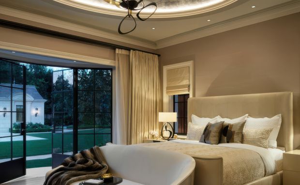 A refined, warm-toned bedroom with a large upholstered bed dressed in layered neutral bedding and decorative cushions. Floor-to-ceiling black-framed glazing opens toward a landscaped garden, while full cream curtains, a curved upholstered bench, bedside lighting, and detailed ceiling mouldings create an elegant, luxurious atmosphere. A sculptural contemporary pendant hangs from a decorative recessed ceiling feature.