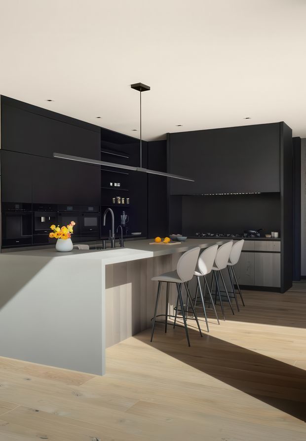 A sophisticated contemporary kitchen with extensive matte-black full-height cabinetry and integrated appliances, a large pale island with a light stone benchtop, and four white sculptural bar stools. A dark linear pendant light hangs over the island, while warm timber flooring and strong sunlight add contrast to the otherwise monochromatic scheme.