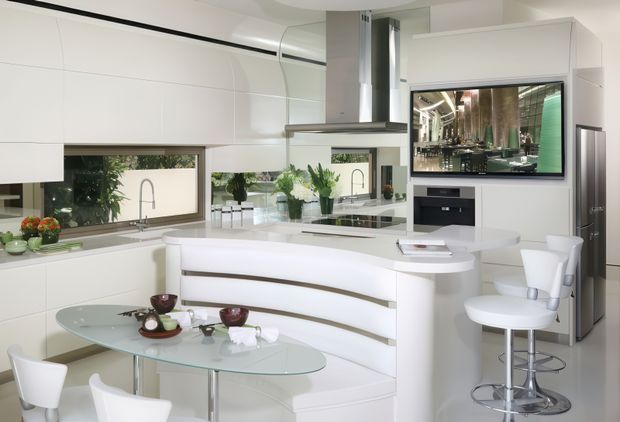 Bright, highly streamlined white kitchen with a curved island and integrated curved banquette seating. The space features handleless white cabinetry, a reflective glass or mirror backsplash, stainless-steel appliances and rangehood, a built-in television, and a glass dining table with white seating. Large windows provide garden views, creating a clean, contemporary open-plan kitchen and dining setting.