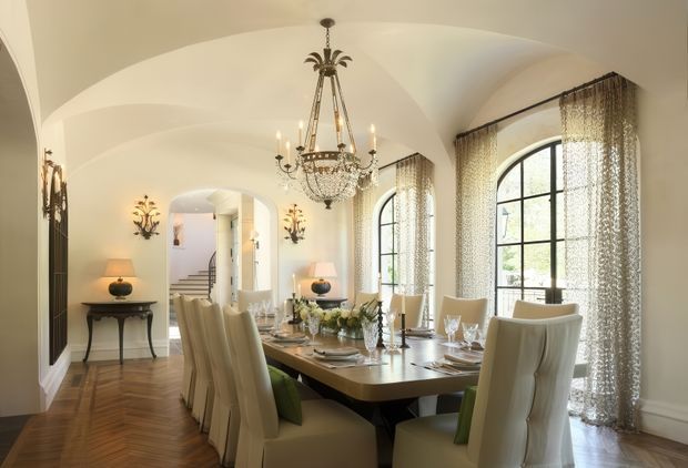 Elegant formal dining room with a long rectangular dining table surrounded by upholstered high-back chairs, set with glassware, plates, candles, and floral arrangements. The room features a dramatic crystal chandelier, arched black-framed windows with sheer patterned curtains, curved vaulted ceiling forms, herringbone timber flooring, ornate wall sconces, and a warm neutral palette. An entry hall and staircase are visible beyond the dining area.