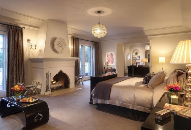 A refined bedroom with a large upholstered bed, fireplace with ornate mantel, plush carpet, tall curtained windows, bedside tables, decorative lighting, and a small workspace or vanity area in the background. The room uses warm cream, taupe, brown, and dark wood tones with traditional detailing and luxurious furnishings.