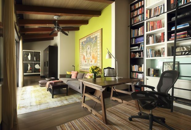 A warm, eclectic open-plan living room and home office with exposed dark timber ceiling beams, a wood-lined ceiling, built-in black-and-white bookcases filled with books, a timber worktable, and an ergonomic office chair. The living area includes a sofa, ottomans, patterned rugs, artwork, a floor lamp, and a ceiling fan. Bright lime-green and neutral walls contrast with the rich timber finishes, creating a comfortable, characterful interior.