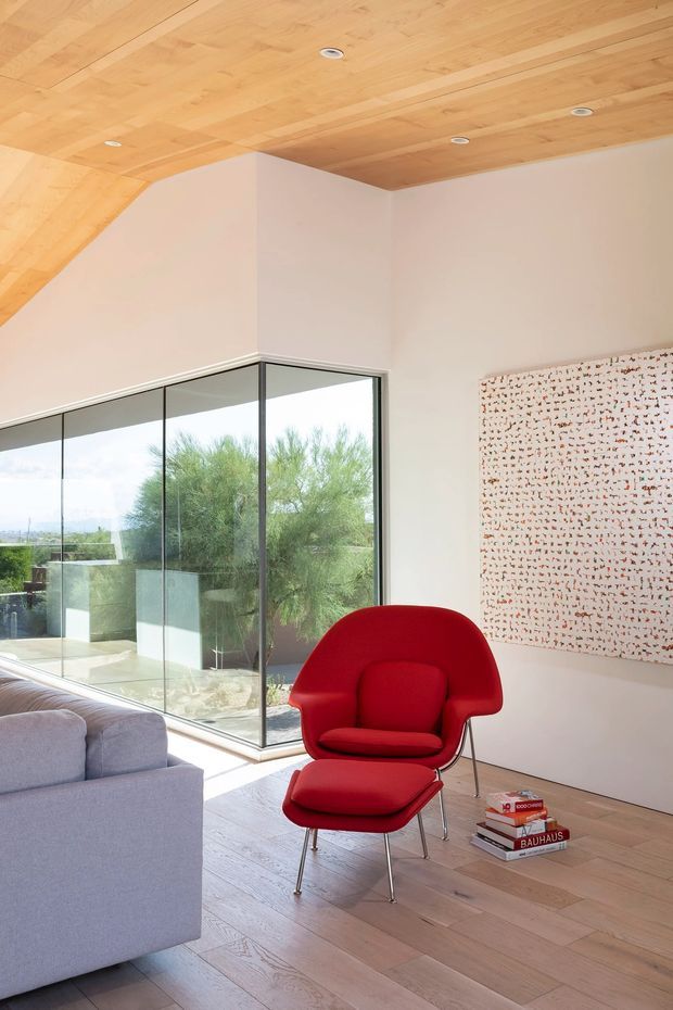 Bright contemporary living room with pale timber flooring and a matching timber-lined sloped ceiling. Full-height corner glazing opens the room visually to an outdoor terrace and greenery. A vivid red sculptural lounge chair and ottoman provide a strong accent beside a light grey sofa, while a large abstract patterned artwork hangs on the white wall. Recessed ceiling lights and minimal detailing create a clean, airy interior.