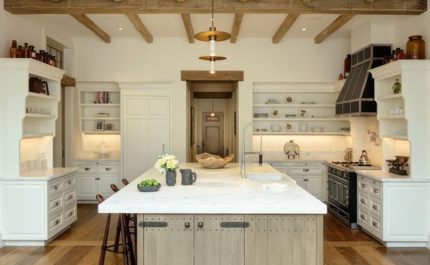 A bright, spacious traditional-style kitchen with a large central island, extensive white built-in cabinetry, open shelving, integrated display niches, and a range cooker beneath a dark angled hood. Exposed timber ceiling beams, warm timber flooring, aged metal hardware, and a timber-framed doorway add rustic character. The island includes seating for several people and a pale stone benchtop, while layered pendant lighting and under-cabinet lighting create a warm, inviting atmosphere.