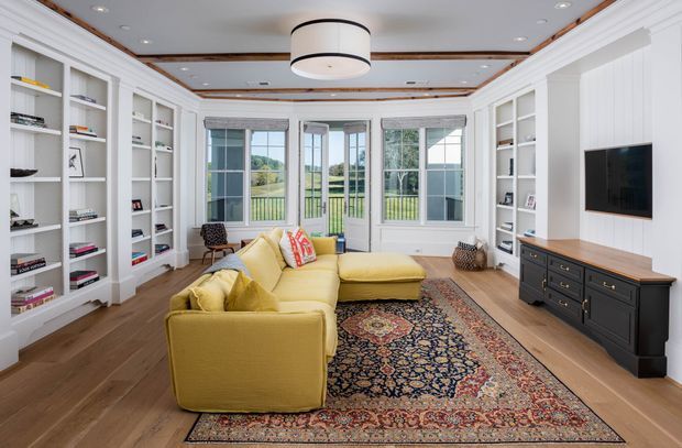 Bright, traditionally detailed living room with built-in white shelving and cabinetry lining both sides, a mustard-yellow sectional sofa, patterned area rug, timber flooring, and a wall-mounted television above a dark built-in media console. Tall glazed doors and windows open to a green outdoor view, while the ceiling features recessed lighting, decorative timber trim, and a large circular pendant light.