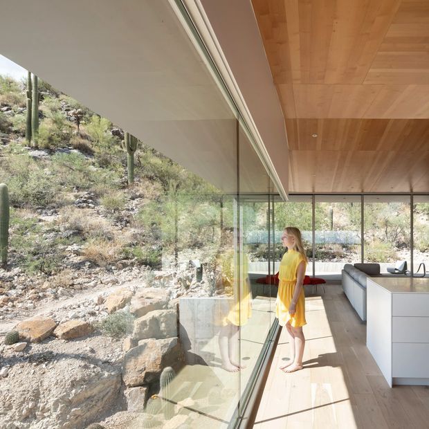 A bright, minimalist open-plan interior with full-height glass glazing overlooking a rocky, cactus-dotted desert hillside. The room features a warm timber-lined ceiling, pale timber flooring, a sleek white kitchen island with integrated cabinetry, and a white sectional sofa. Strong reflections in the glazing create a layered connection between the interior and rugged landscape.