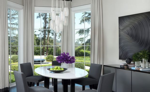 Bright contemporary dining area with a round white dining table and dark upholstered chairs, positioned in front of tall multi-pane corner windows with sheer curtains. A cluster of glass pendant lights hangs above the table, while a dark built-in sideboard and abstract monochrome artwork occupy the right wall. Landscaped greenery is visible outside.