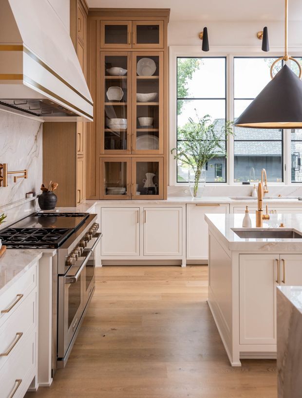 Bright, refined kitchen interior with a large central island, white inset-panel cabinetry, warm timber flooring, a tall glass-fronted timber display cabinet, marble-look splashback and benchtops, brass hardware and tapware, and a black statement pendant light. Large divided windows provide abundant natural light and overlook neighbouring buildings.