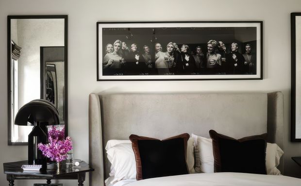 A refined bedroom vignette featuring a tall upholstered bedhead in a pale neutral fabric, crisp white bedding, and dark velvet accent cushions. Black-framed monochrome artwork hangs above the bed, with a large black-framed mirror and a small bedside table with a black lamp and flowers to the left. The room has a balanced, sophisticated monochrome palette with subtle traditional detailing.