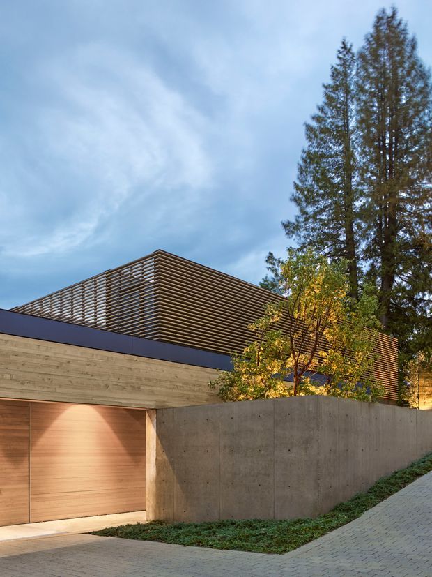 Street-level exterior view of a contemporary residence, featuring a warm timber-clad garage door and horizontal timber wall, a tall raw concrete boundary wall, and a prominent upper volume screened with closely spaced horizontal timber louvres. Mature trees surround the building, and a paved sloping driveway occupies the foreground.