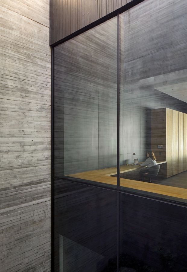 A dramatic architectural interior view through full-height dark-framed glazing, showing a concrete-lined space with a warm timber floor, vertical timber wall panelling, and a seated person at a desk or table. The composition emphasises raw concrete, black window framing, strong vertical lines, and reflected interior surfaces.