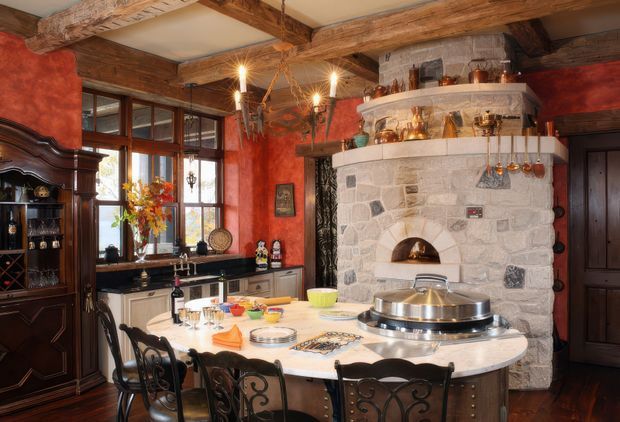 A richly detailed rustic kitchen and dining space with a large central island, ornate black metal seating, dark wood cabinetry, and a substantial pale stone fireplace with a built-in oven or hearth. Exposed rough-hewn timber beams cross the ceiling, while warm red textured walls, leaded or mullioned timber-framed windows, copper cookware, and traditional decorative joinery create a historic European farmhouse feel.