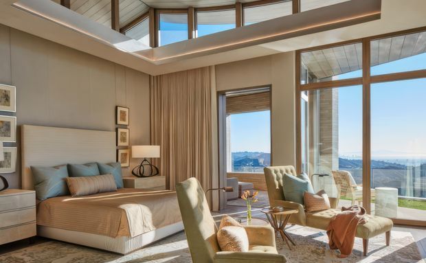 A sophisticated bedroom sitting area with a large upholstered bed, bedside tables, lounge chairs, and a small round coffee table. Floor-to-ceiling glazing and clerestory windows provide expansive views, while timber-framed glazing, vertical wall drapery, warm neutral finishes, and a high ceiling create an airy, refined interior.