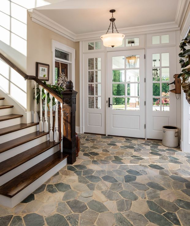 A bright, traditional entry foyer with a dark-stained timber staircase, ornate turned balusters and carved newel post, white painted trim and panelled French-style entry doors with extensive glazing. The floor is finished in irregular natural stone flagging, and a classic pendant light hangs from a coffered and heavily moulded ceiling. Garden greenery is visible through the doors.