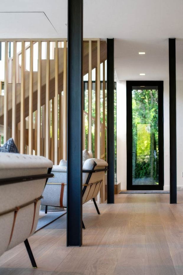 A bright contemporary interior hallway or entry zone with pale timber flooring, white walls and ceiling, black-framed glazing and doors, and a timber slatted screen enclosing a staircase. Upholstered lounge chairs are visible beside the circulation space, with greenery seen through the glazed rear door.