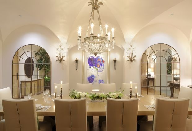 Elegant formal dining room with a long light-toned dining table and upholstered dining chairs arranged beneath an ornate crystal chandelier. The room has warm cream walls, arched vaulted ceiling forms, large black-framed arched glazed openings, decorative wall sconces, candles, floral arrangements, and a colourful artwork centred on the rear wall. The overall ambience is refined, warm, and luxurious.