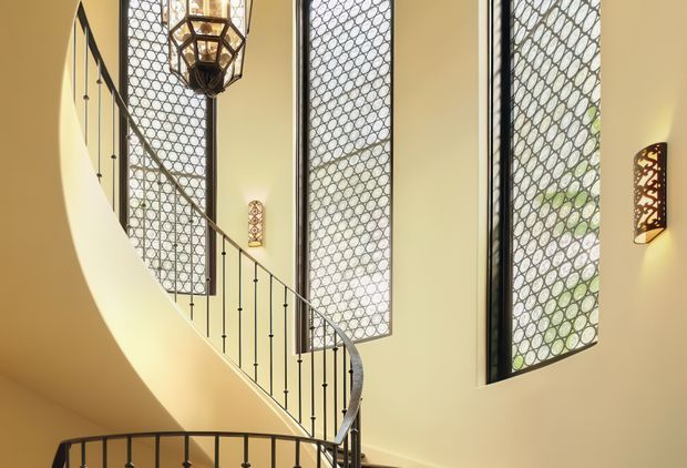 A tall, warmly lit stairway with a curved or spiral form, slender dark metal balustrade, and expansive vertical windows fitted with decorative geometric metal screens. A large hanging lantern and matching patterned wall sconces add an ornate, refined character. The space appears to have a high ceiling and strong natural light filtering through the screens.
