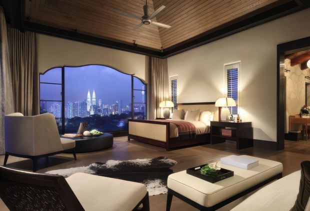 A luxurious contemporary bedroom with a large arched window overlooking a brightly lit city skyline at dusk. The room features a timber-lined ceiling with exposed dark trim, dark flooring, a large bed, upholstered lounge chairs, a low coffee table, bedside tables, floor-length curtains, and warm ambient lighting. An open doorway reveals an adjacent bathroom or dressing area.