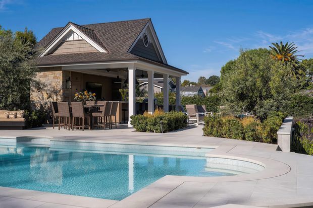 A bright outdoor residential pool area beside a covered pavilion or veranda. The scene features a curved swimming pool with pale paving, landscaped garden beds, outdoor lounge chairs, and a dining/bar setting under a gabled, shingle-clad roof supported by white columns. The architecture has a traditional suburban character with prominent gables and detailed trim.