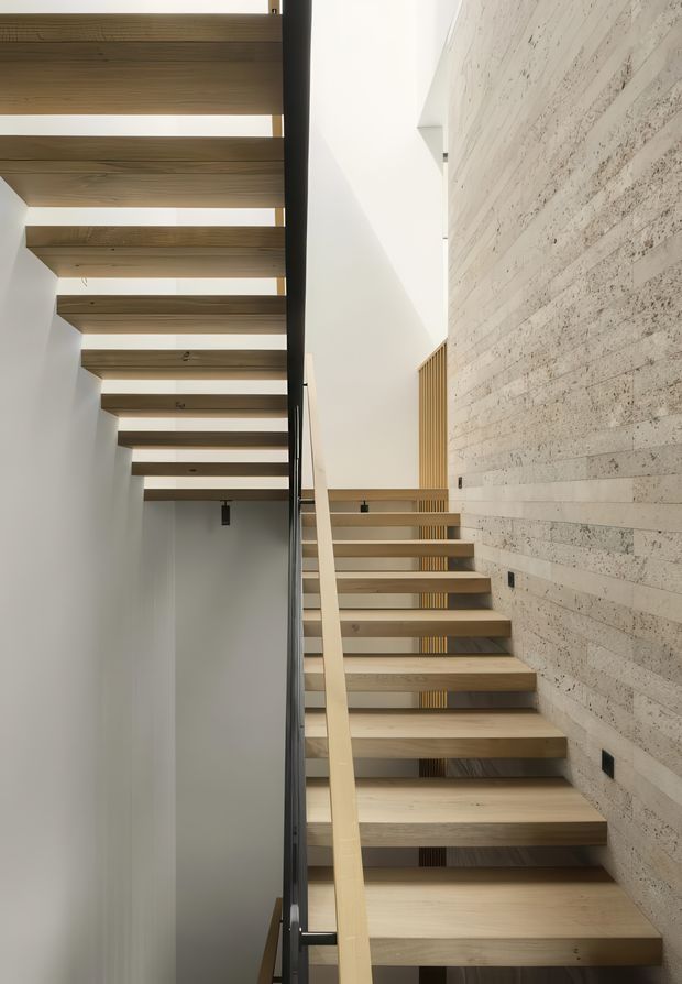 Interior view of a contemporary stairway rising alongside a textured pale masonry feature wall. The open-riser staircase and overhead floating timber beams create a strong linear architectural composition, with a slim dark metal balustrade and warm timber detailing. A bright void above and beside the stairs suggests a tall ceiling or vertical circulation space.