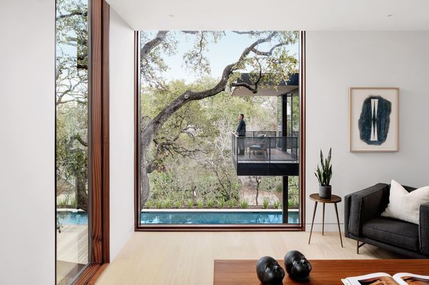 A refined contemporary living room with pale timber flooring, white walls and expansive floor-to-ceiling glazing framing mature trees, a swimming pool and an elevated outdoor balcony. The room includes a dark upholstered sofa, sculptural coffee table, potted plant and framed artwork. Warm timber window framing and the strong indoor-outdoor connection define the space.