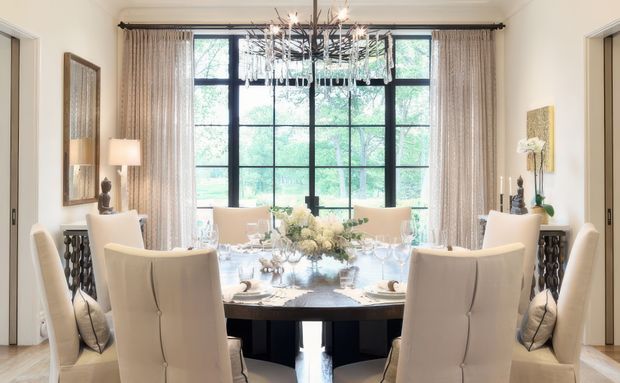 Elegant formal dining room centred on a large rectangular dining table with upholstered high-back chairs, refined table settings, a floral arrangement, and a dramatic branching chandelier. Full-height black-framed glazing with sheer curtains fills the rear wall, while light neutral finishes and dark accent furniture create a bright, sophisticated interior.