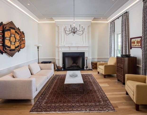 A refined living room with a large ornate white fireplace and mantel as the focal point. The room features layered ceiling coffers with recessed lighting, white wall panelling and cornices, tall patterned curtains, pale upholstered sofas and armchairs, a marble or stone coffee table, a vintage Persian-style rug, a sculptural chandelier, timber flooring, and mid-century-inspired timber cabinetry. Decorative artwork and a television are visible along the walls.