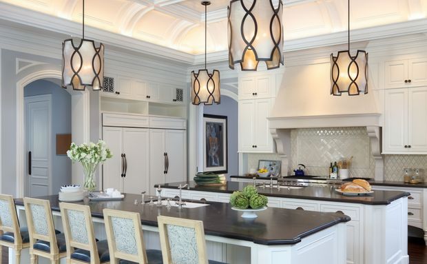 A bright, spacious traditional kitchen with extensive white paneled cabinetry, a large dark island with seating, integrated appliances, a pale herringbone tile backsplash, and a substantial decorative rangehood surround. Three oversized pendant lights with black looping metalwork hang above the island and dining area. Arched doorways, ornate ceiling detailing, and layered trim create a refined, classic interior.