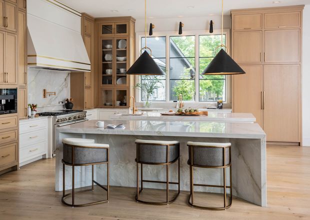 Bright, refined kitchen interior with a large marble-clad island and seating for three. The room features warm wood cabinetry, integrated appliances, a sculptural white rangehood, brass-accented hardware and trims, black-and-brass pendant lights, timber flooring, and large windows overlooking greenery.