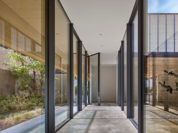 A minimalist, light-filled glazed passageway with full-height dark metal-framed glass walls and doors on both sides. The corridor has a smooth pale concrete floor, a simple white ceiling with recessed downlights, and views into landscaped courtyards and timber-lined spaces through the glazing. Horizontal external sun-shading screens cast patterned shadows across the floor and walls.