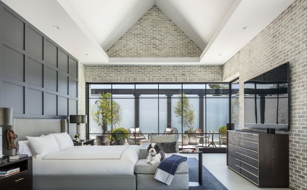 A bright, spacious contemporary bedroom with a pitched vaulted ceiling and exposed light brick feature walls. The room contains a large upholstered bed, a grey ottoman at the foot, dark built-in wall panelling and cabinetry, a television on a timber-toned dresser, and full-height black-framed glazing opening toward a planted outdoor area. A small dog is present on the bed, and the overall palette is neutral with strong black and grey accents.