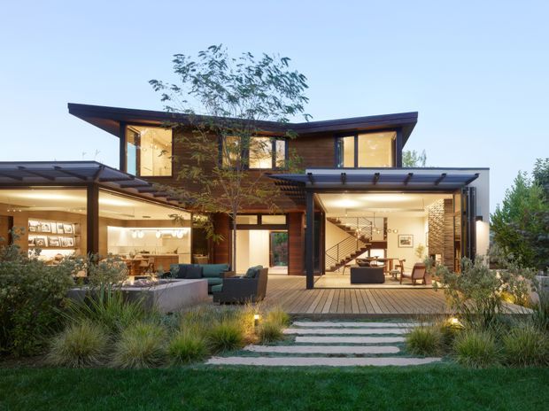 A contemporary two-storey residence viewed from the landscaped front garden at dusk. The home features broad floor-to-ceiling glazing, warm timber cladding, dark metal roof edges and structural framing, covered outdoor living areas, and an illuminated open-plan interior with a visible staircase. A timber deck, stepping-stone path, ornamental grasses, and layered planting connect the house to the garden.