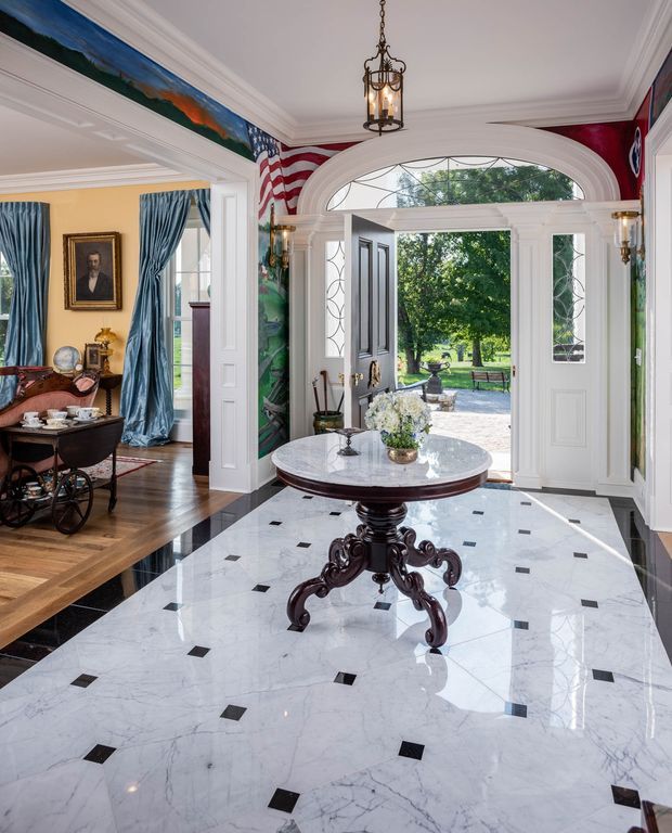Elegant traditional entry foyer with a polished white marble floor patterned with small black square inlays, ornate dark wood round table, white paneled walls and elaborate crown moulding. A large arched glazed front door opens to the garden, flanked by decorative sidelights. A formal sitting room with blue drapery, timber flooring and antique furnishings is visible through the opening to the left. American flag-themed wall artwork and classical lantern lighting add a historic, refined character.