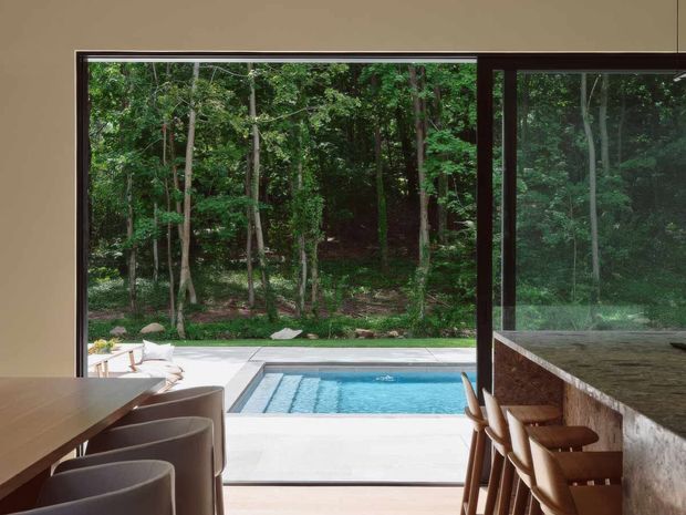 Contemporary indoor-outdoor dining and bar area opening directly onto a rectangular swimming pool and landscaped woodland. Dark-framed floor-to-ceiling glazing creates a strong connection to the forest, while a long timber dining table with upholstered chairs occupies the left side and a stone-faced bar with timber stools lines the right. The space has a restrained, refined palette and abundant natural light.