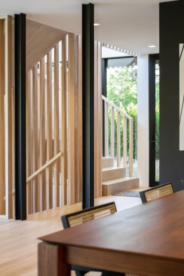 A bright contemporary interior centered on a timber staircase and open circulation area beside a dining space. Vertical timber battens form a balustrade or screening element, contrasting with black-framed glazing and dark vertical wall panels. A large timber dining table and woven-back chairs occupy the foreground, while greenery is visible through the windows. The space has clean lines, abundant natural light, and a warm timber palette.