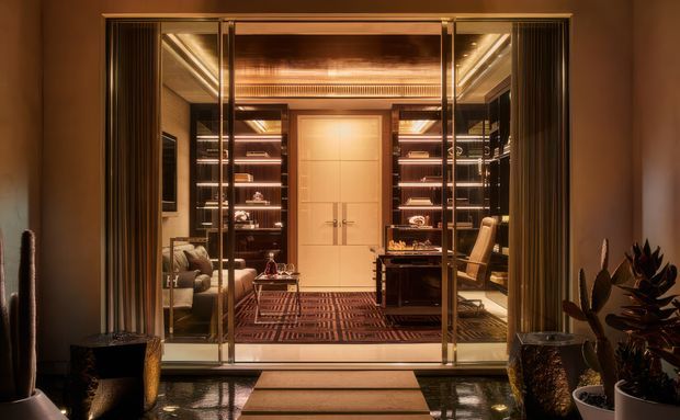 A luxurious, warm-toned interior viewed through full-height glass doors, showing a refined home office or study with extensive illuminated built-in shelving, a central paneled double door, desk and office chair, lounge seating, patterned floor covering, and dark glossy flooring. Symmetrical framing, bronze-toned finishes, and layered lighting create an intimate, high-end atmosphere.