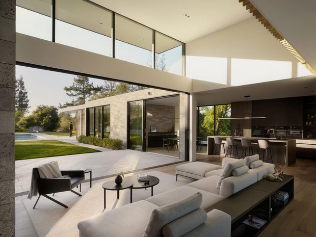 A spacious contemporary open-plan living room with a double-height glazed corner opening onto a landscaped garden and pool. The room contains a large light upholstered sectional sofa, black-framed lounge chair, round coffee tables, and a long dark built-in media/storage unit. A modern kitchen with a dark island and bar seating is visible to the right. The architecture features expansive floor-to-ceiling glazing, a projecting upper-level walkway or mezzanine, clean rectilinear volumes, and a linear timber detail along the ceiling edge.