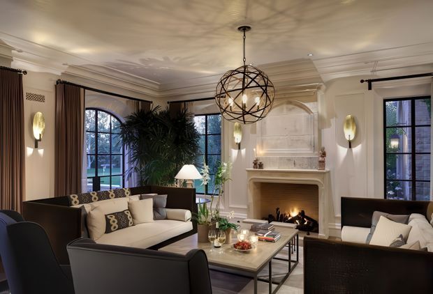 Elegant, warmly lit living room with a classical fireplace, ornate ceiling mouldings, tall black-framed arched and rectangular windows, layered curtains, dark upholstered sofas and armchairs, and a central geometric spherical pendant light. A fire burns in the mantel fireplace, creating a refined and inviting atmosphere.