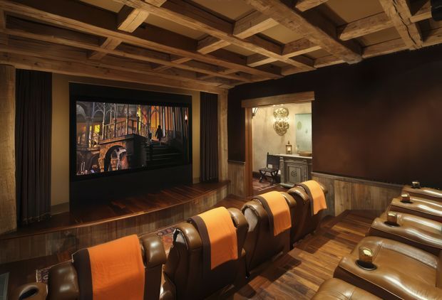 A dark, luxurious home cinema with a large projection screen, several rows of plush brown leather recliner seats draped with orange throws, and extensive warm timber finishes. The ceiling features exposed timber beams and a coffered grid, while a timber-lined wall and doorway lead to a small adjoining area with cabinetry and decorative lighting.