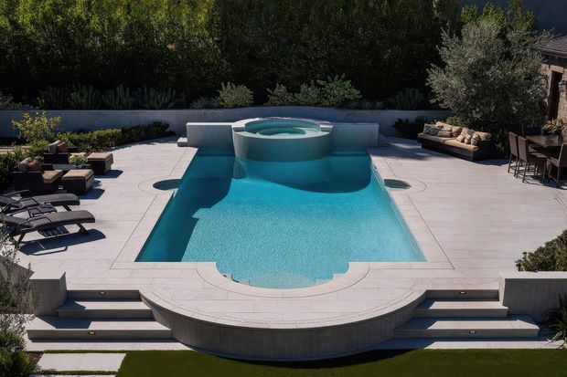 A luxurious outdoor pool area viewed from above, featuring a large turquoise swimming pool with a raised circular spa, sculptural curved concrete pool edge, broad pale stone paving, integrated tiered steps, sun loungers, built-in seating, and landscaped screening with mature greenery. The composition is symmetrical and refined, with strong geometric and curved forms.