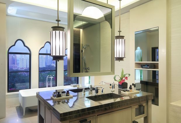 Luxurious contemporary bathroom with a large central double vanity featuring a dark polished stone benchtop, integrated basins, metallic fixtures, and abundant built-in storage. Tall arched windows, a freestanding bathtub, large framed mirror, glass shower enclosure, fluted wall detailing, and pendant lighting create a refined hotel-like interior. The room is bright with warm neutral finishes and dark contrasting joinery.