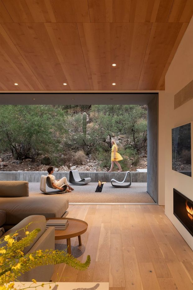 A warm, contemporary living room opens through a large glazed or retractable opening to an outdoor seating terrace and natural rocky landscape. The interior features a pale timber floor and matching timber-panelled ceiling with recessed downlights, a large neutral sectional sofa, sculptural lounge chairs, a round timber coffee table, and a built-in dark fireplace on the right wall. A small outdoor fire pit and additional seating are visible beyond the opening.