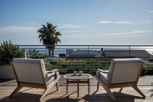 A sunlit elevated outdoor terrace or balcony with two cushioned timber-framed lounge chairs and a small round table, overlooking rooftops and the ocean beyond. Horizontal metal railings, timber decking, planter greenery, and a palm tree create a relaxed coastal setting.