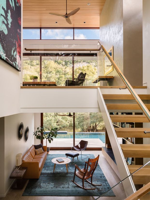 A bright double-height interior showing an open stairway and mezzanine overlooking a living room. The space features expansive timber-framed glazing, a warm timber slatted ceiling, floating timber stair treads with glass balustrades, and mid-century-style leather seating around a small round coffee table. A swimming pool and leafy landscape are visible outside through the windows.