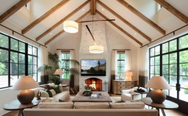 A bright, spacious living room with a vaulted white ceiling and exposed timber rafters. Large black-framed windows line both sides, overlooking greenery. The room features a neutral sectional sofa, armchairs, round side tables with ceramic lamps, a central coffee table, indoor palms, a painted brick fireplace with a television above it, and warm timber furniture. The overall design combines refined traditional detailing with relaxed contemporary furnishings.