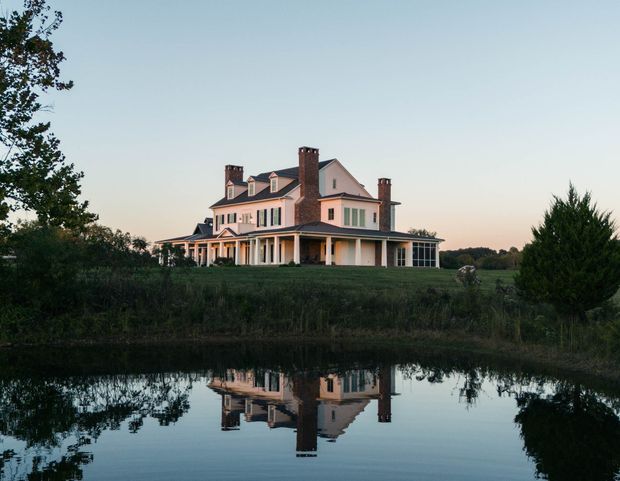 A large stately multi-storey country house viewed across a reflective pond at dusk. The pale pink-beige rendered facade has dark shutters, multiple steep gabled roof sections, several tall brick chimneys, and an expansive covered veranda supported by evenly spaced white columns. The home sits in a spacious rural landscape with mature trees and lawn, while the calm water provides a clear reflection.