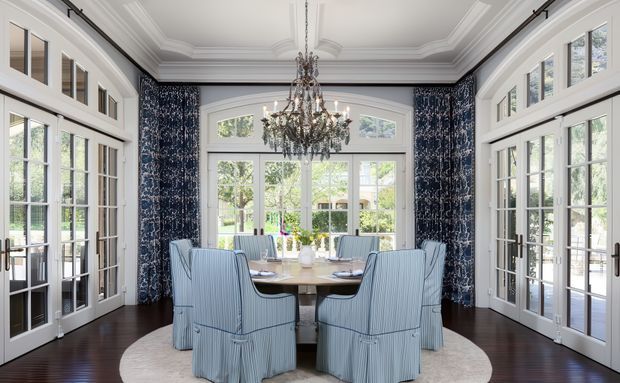 Elegant formal dining room with a large round dining table surrounded by slipcovered blue upholstered chairs. The room features extensive white French-style multi-pane glazing and glazed doors, deep navy patterned drapery, dark timber flooring, an ornate crystal chandelier, and a highly detailed coffered and moulded ceiling. Abundant daylight and garden views create a refined, bright atmosphere.